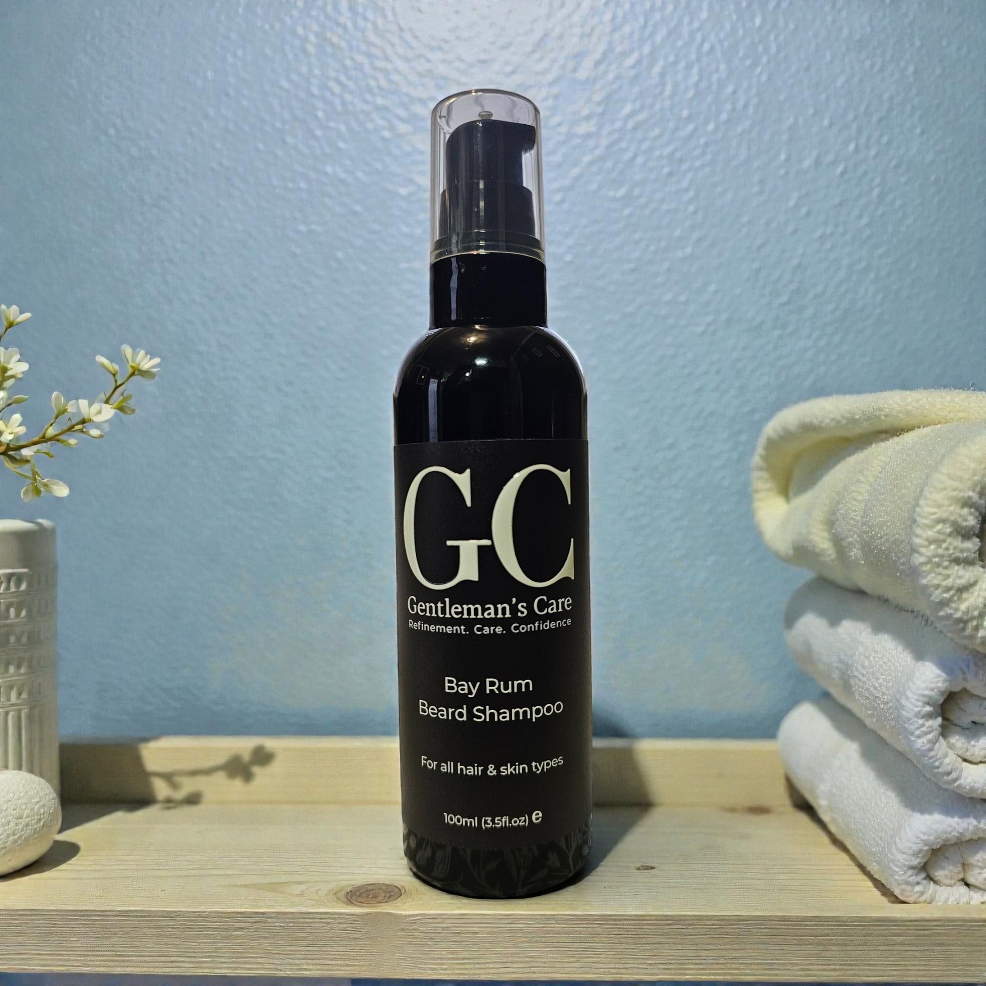Bottle of Gentleman's Care Bay Rum Beard Shampoo on a wooden surface with towels and a vase in the background.