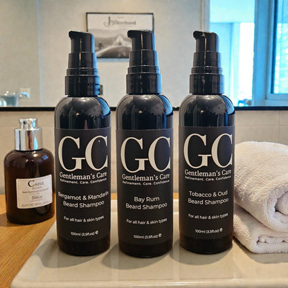 Three bottles of Gentleman's Care beard shampoo on a bathroom counter with a towel and another bottle in the background.