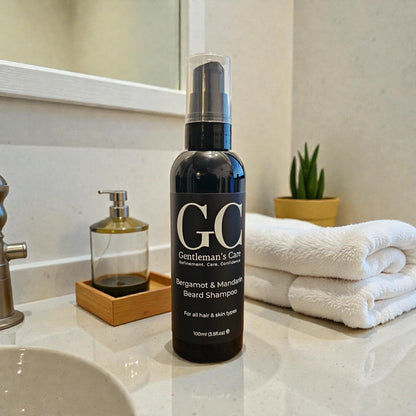 Bottle of 'GO Gentleman's Care' beard shampoo on a bathroom counter with towels and a plant in the background.