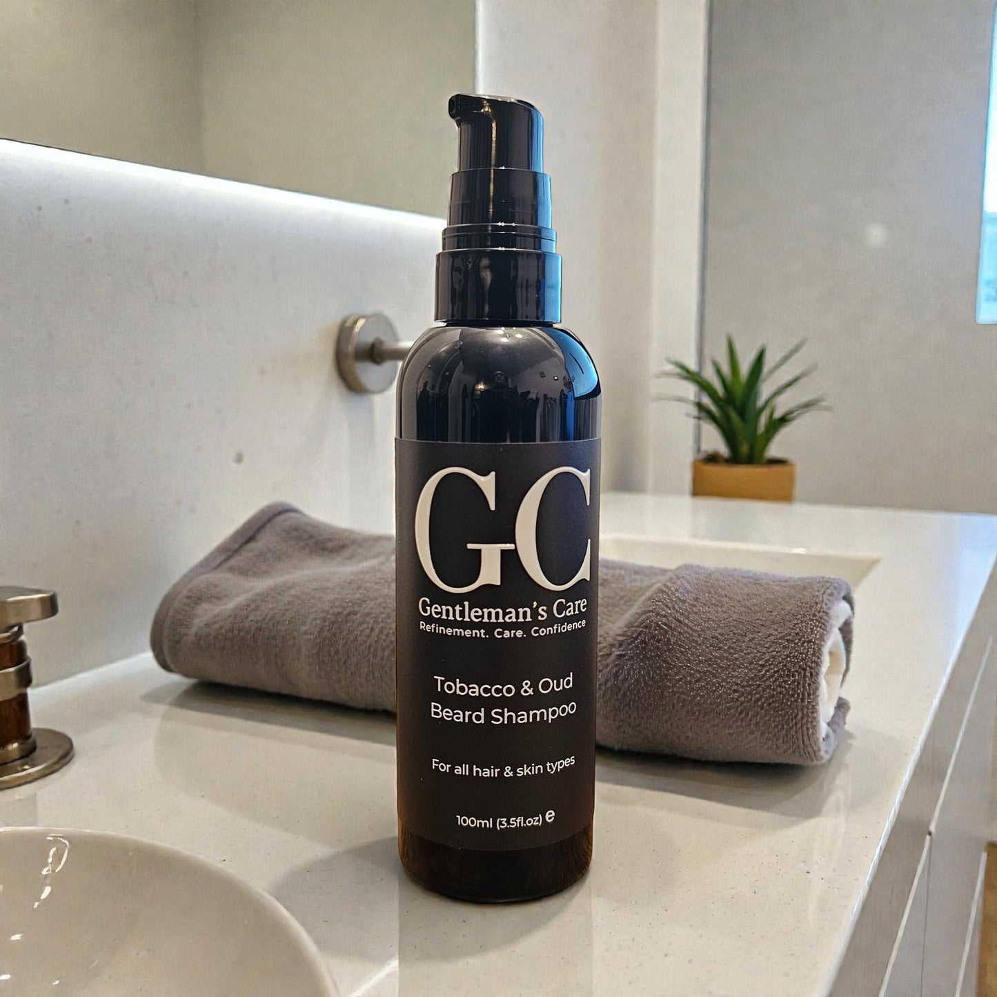 Bottle of Gentleman's Care Tobacco & Oud Beard Shampoo on a bathroom counter with towels and a plant in the background.
